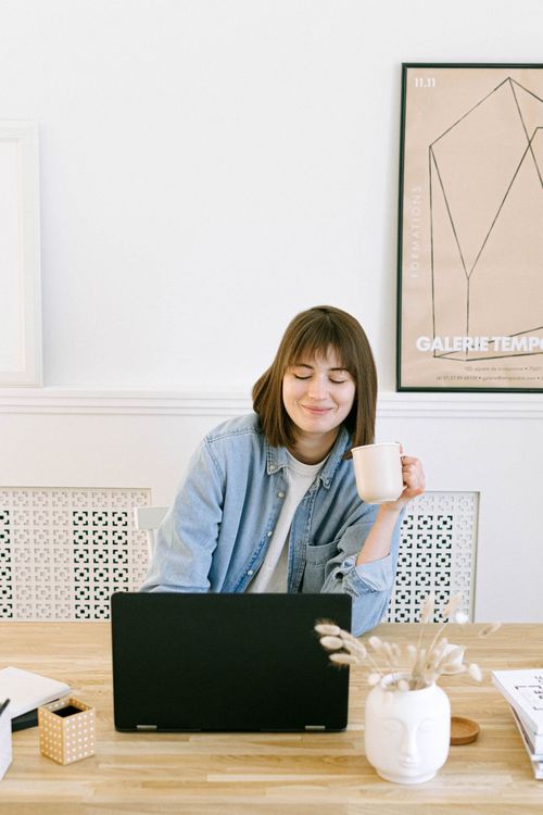 A woman with a coffee cup seated at a laptop in a bright room